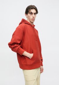 Κόκκινο βαμβακερό φούτερ με κουκούλα, μπροστινή τσέπη, ρυθμιζόμενο κορδόνι στην κουκούλα και ριμπ μανσέτες. Το μοντέλο φοράει ανοιχτό μπεζ παντελόνια cargo.