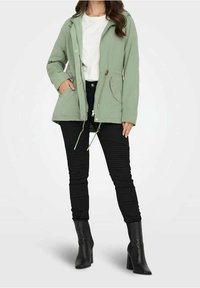 Jasnozielona kurtka na zamek z kapturem, z dwoma przednimi kieszeniami, ściągaczem w pasie oraz czarne spodnie z prążkowanego materiału, połączone z czarnymi butami na obcasie.