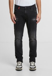 Jeans de mezclilla negra con corte ajustado, con un ligero desgaste, cinco bolsillos y un acento rojo en la cintura. Combinados con zapatillas negras y blancas.