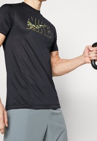 Camiseta atlética negra con un logo de camuflaje en el pecho, fabricada con un material suave y elástico. Mangas cortas, diseño ajustado.