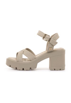 Beige sandal med blokhæl, tyk sål med mønster, krydsede stropper og ankelrem med guldbøjle på hvid baggrund.