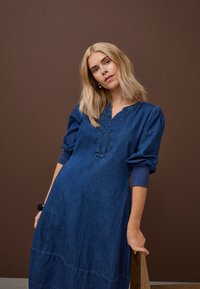 Blondína v modrých džínsových šatách s dlhým rukávom opretá o drevenú stoličku, na jednofarebnom hnedom pozadí.