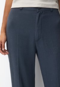 Kobieta ubrana w granatowe dopasowane spodnie oraz beżowy bawełniany top, który jest włożony do środka, stoi z lewą ręką luźno wzdłuż ciała.