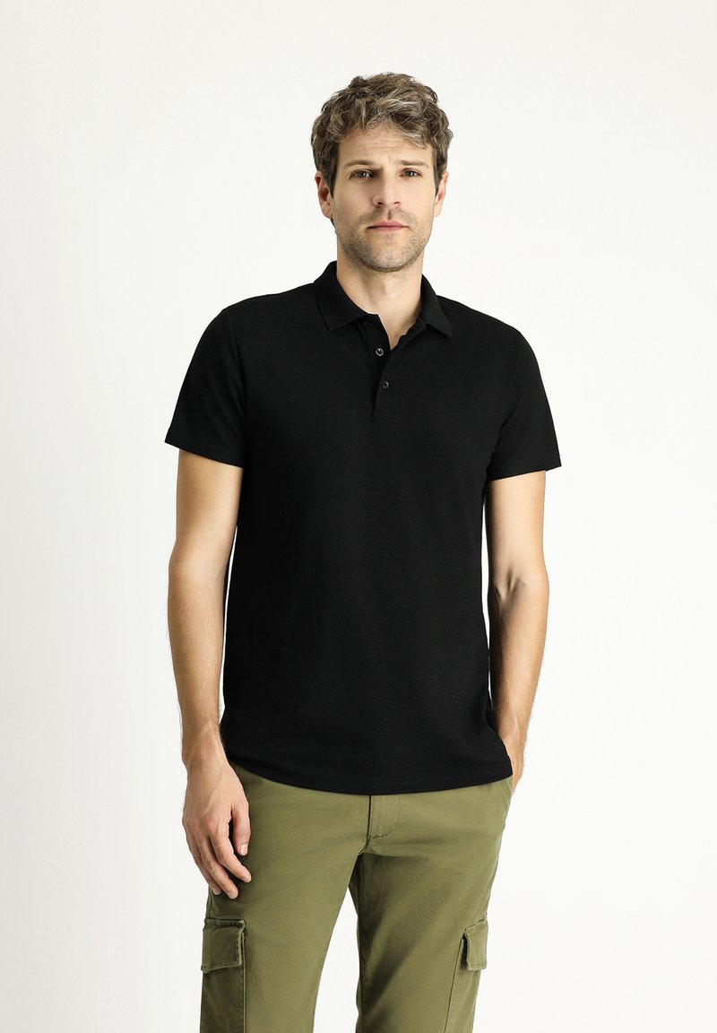 Camisa polo negra de manga corta, con tapeta de tres botones, textura suave, combinada con pantalones cargo verde olivo. Diseño simple y clásico.