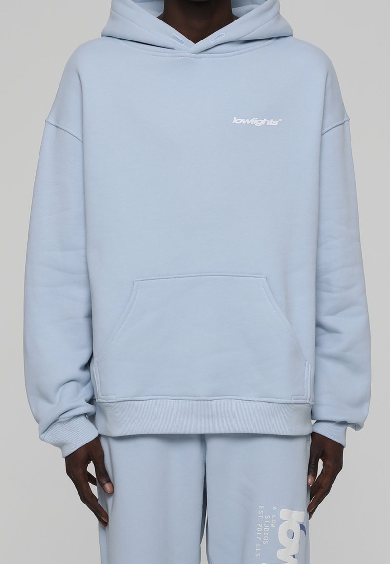 Sudadera azul claro con un bolsillo frontal y puños acanalados. Presenta un pequeño logotipo blanco en el pecho y pantalones de chándal a juego con un diseño de texto.