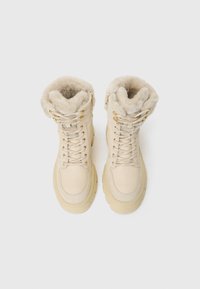 Beige, cipőfűzős csizmák bolyhos krémszínű szőrme gallérral, arany hardware részletekkel és texturált talpdesignnal. Puha bőr és velúr anyagok.