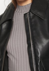 Schwarze Lederjacke mit Reverskragen und silbernem Reißverschluss, darüber ein graues geripptes Oberteil mit hohem Ausschnitt und vertikalen Streifen.