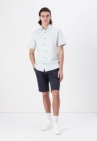 Chemise à manches courtes bleu clair avec des boutons noirs, accompagnée de shorts bleu marine foncé et de baskets blanches. Texture de tissu lisse.