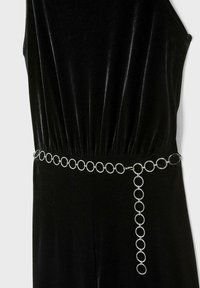 Schwarzer Samt-Jumpsuit mit gerafftem Bund und einem silbernen runden Kettengürtel, der das Design mit einem markanten hängenden Detail akzentuiert.