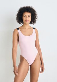 Maillot de bain une pièce en rose doux avec des bretelles noires, présentant un décolleté rond et des ouvertures de jambe hautes, fabriqué en tissu lisse.