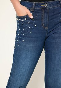 Dunkelblaue Denim-Jeans im Slim Fit mit Perlenverzierungen an den Vordertaschen und Seitennähten. Auffällige gelbe Steppnähte.