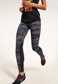 Černé sportovní legíny s texturovaným vířivým bílým vzorem. Mají obepínající střih a síťované ventilační panely po stranách.