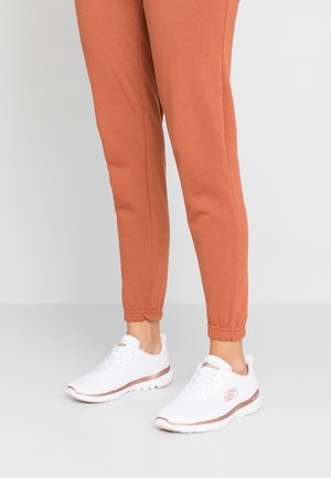 Persona che indossa scarpe sportive bianche con dettagli in oro rosa e pantaloni jogger color ruggine, in piedi su un pavimento neutro.