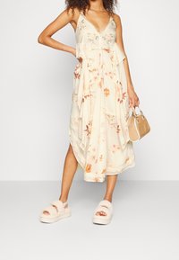 Blommig midiklänning i krämfärgat med volangdetaljer och spetskant. Har spagettiband och en svepande design. Matchas med lätta, fluffiga sandaler.