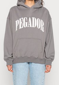 Grauer Kapuzenpullover mit der Aufschrift "PEGADOR" in Weiß. Verfügt über eine Kängurutasche und gerippte Bündchen. Der Stoff wirkt weich und strukturiert.