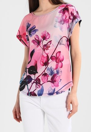 Kobieta ubrana w różową bluzkę z krótkim rękawem z dużym fioletowo-magentowym motywem kwiatowym, połączoną z białymi spodniami, stojąca na jednolitym tle.