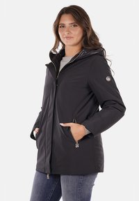 Cappotto impermeabile nero con cappuccio, tasche con zip e una texture liscia. Presenta un accento con logo sulla manica per un tocco di dettaglio in più.