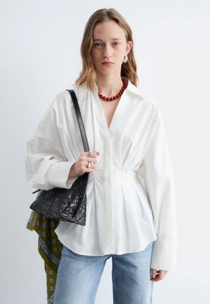 Ung kvinde iført en løs hvid bluse, lyseblå jeans, rødt perlekrave, der holder en sort tekstureret håndtaske med et grønt mønstret tørklæde.