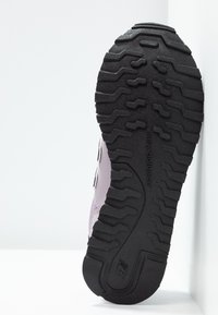Czarna gumowa podeszwa z głębokim wzorem bieżnika na sportowym bucie New Balance, ustawionym pod kątem na białym narożnym tle.