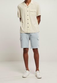 Chemise à manches courtes, couleur beige clair, avec un poche poitrine et des boutons noirs, associée à un short cargo bleu clair et des baskets blanches.
