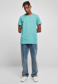 Tricou din bumbac turcoaz cu gât rotund, mâneci scurte, asortat cu blugi din denim deschis, deteriorați, și adidași albi.