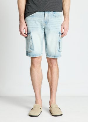 Uomo che indossa pantaloncini cargo in denim azzurro chiaro e scarpe slip-on beige, in piedi su uno sfondo bianco semplice.