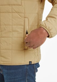Beige quilted jacket with elastic cuffs and a zipper pocket on the front. Fabric appears smooth with a slight sheen. Detailed stitching visible.