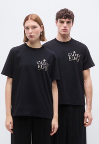 Μαύρα βαμβακερά μπλουζάκια με κοντά μανίκια, που διαθέτουν μικρό λευκό και χρυσό λογότυπο «CALVIN KLEIN» στο αριστερό στήθος. Και τα δύο μοντέλα φορούν μαύρο παντελόνι.