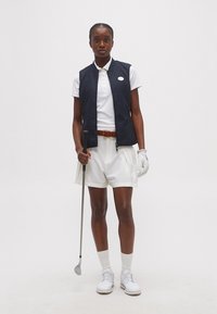 Γυναίκα που φοράει λευκή γκολφ ενδυμασία και μαύρο γιλέκο, κρατώντας ένα γκολφ ρόπαλο, στέκεται μπροστά σε ένα απλό λευκό φόντο.