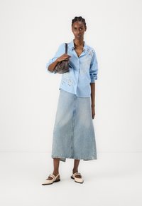 Camisa de botão azul-claro com riscas e bordado floral, combinada com uma saia longa de ganga e mocassins creme. Pequena bolsa castanha.
