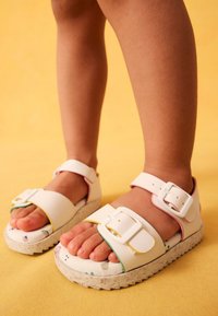 Weiße Sandalen mit verstellbaren Riemen, strukturierter Sohle und bunten Akzenten. Die Innensohle zeigt ein verspieltes Muster auf einer leichten Plateausohle.