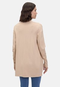 Beige cardigan van zacht breisel, met een lange snit, afgeronde zoom en knoopaccenten op de mouwen. G gedragen over blauwe jeans.