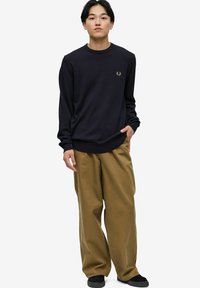 Suéter náutico con cuello redondo y puños acanalados, que presenta un logo dorado. Combinado con pantalones khaki sueltos y zapatos negros. Diseño simple.