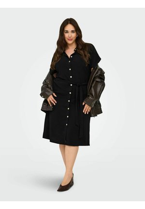 Donna con abito nero abbottonato con cintura in vita indossa una giacca di pelle marrone scuro drappeggiata sulle spalle e scarpe abbinate con punta appuntita.