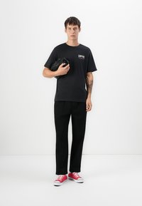 Ung mand i sort t-shirt med Empire-logo, sorte bukser, røde sneakers, holder en sammenfoldet sort jakke, stående mod hvid baggrund.