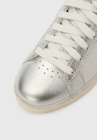 Ασημί δερμάτινο αθλητικό παπούτσι με λευκά κορδόνια, διάτρητη λεπτομέρεια κοντά στα δάχτυλα και υφασμένο μπεζ καουτσούκ πάτο σε λευκό φόντο.