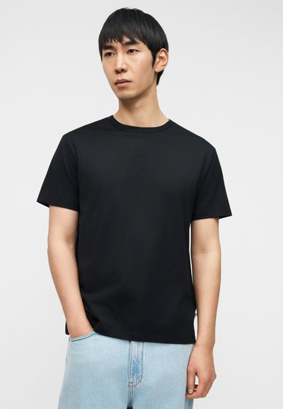 T-shirt noir à col ras du cou en tissu doux, manches courtes et coupe décontractée, associé à un jean bleu clair. Design simple sans motifs visibles.