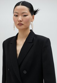 Svart dubbelknäppt kavaj med spetsiga slag, med en slät tygstruktur och en enda svart knappprydnad. Guld geometriska örhängen synliga.