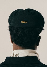 Persoon met kort, krullend haar draagt een zwarte pet met "Blkvio" geborduurd in goud, ringoorbellen, een patroon van een witte nektekel, en een zwarte trui.