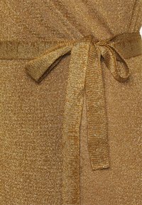 Gros plan d'un tissu métallique doré avec un tricot texturé et un nœud noué en tissu identique autour de la taille.