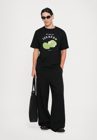 Man wearing black sunglasses, black t-shirt with iceberg lettuce graphic, wide black pants, and holding a black tote bag.