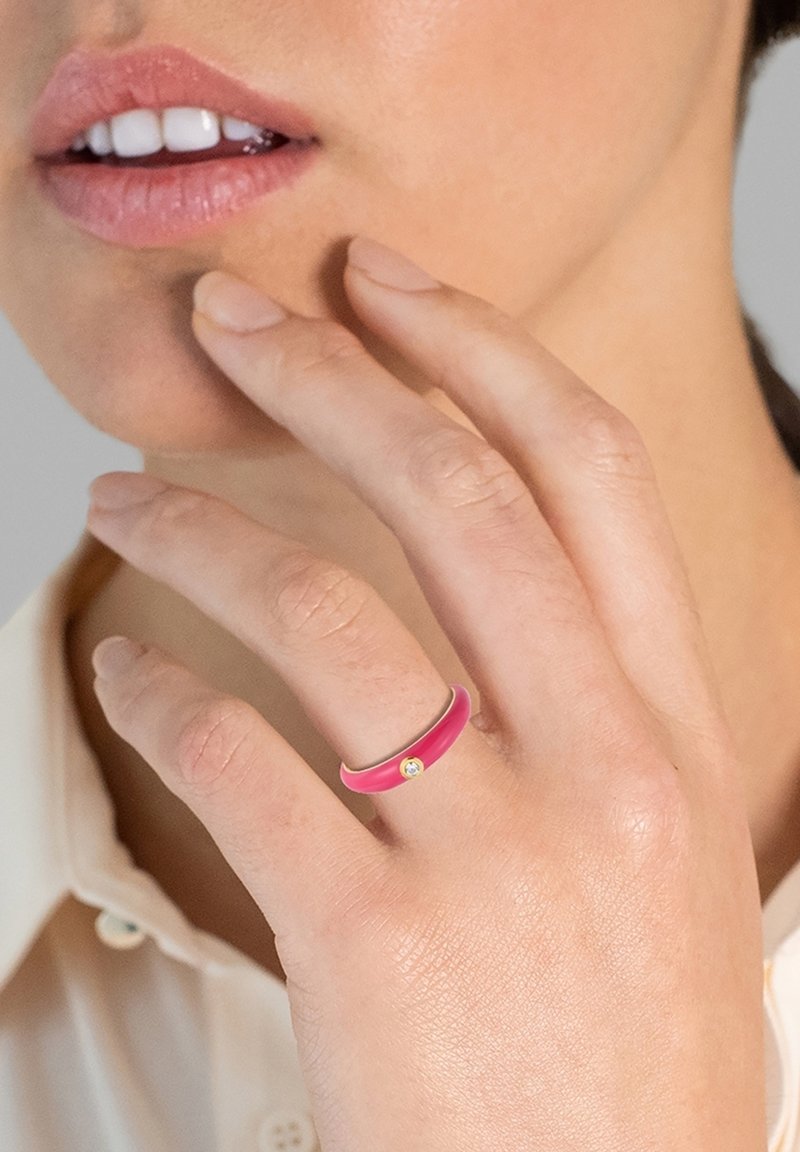 Rózsaszín gyűrű sima felülettel és egy kis drágakő hangsúllyal. Kézen viselve, világos színű inggel. A bőr textúrája látható.