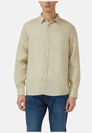 Mężczyzna ubrany w beżową koszulę na długi rękaw z guzikami oraz niebieskie dżinsy, stojący na jednolitym tle.