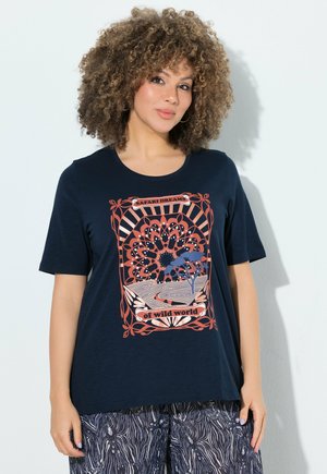 Femme aux cheveux bouclés portant un t-shirt bleu marine avec l’inscription « Safari Dreams of wild world » et un pantalon foncé à motifs, debout contre un mur blanc.