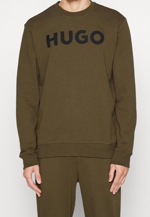 Henkilö, jolla on yllään oliivinvihreä pitkähihaisuus collegepaita ja siihen sopivat housut, joissa on musta "HUGO"-logo rinnassa.