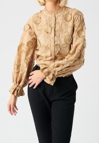Beige blouse with floral embroidery, button closure, ruffled collar and cuffs, long sleeves, and a textured fabric, paired with black pants.