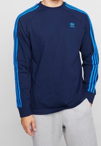 Hombre vestido con una camiseta de manga larga Adidas de color azul marino con franjas azules brillantes en las mangas y pantalones de chándal grises, con una mano en el bolsillo.