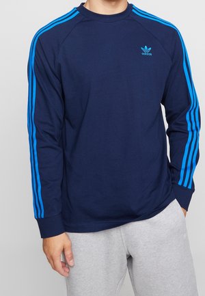 Man in een marineblauwe Adidas lange mouwen shirt met felblauwe strepen op de mouwen en grijze sweatpants, hand in de zak.
