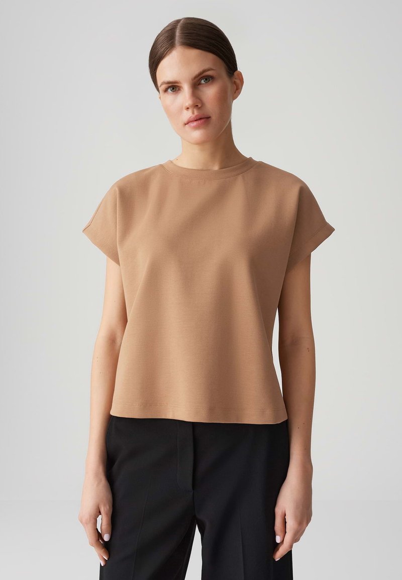 Top beige in cotone misto a maniche corte con scollatura rotonda, vestibilità comoda e texture liscia, abbinato a pantaloni neri.