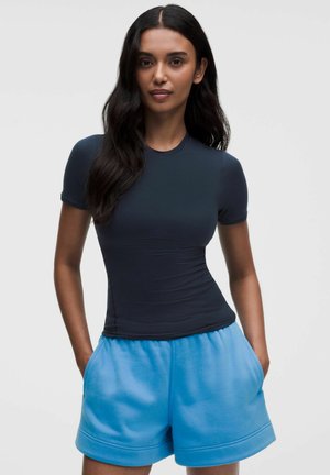 Vrouw met lang donker haar staat met handen in zakken en draagt een aansluitend marineblauw T-shirt met korte mouwen en losse blauwe shorts tegen een witte achtergrond.